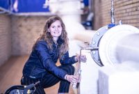 Smiling woman fills a bottle from an industrial pipe in a factory setting.